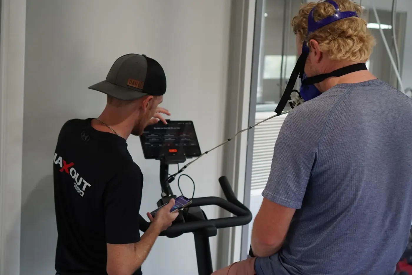 A coach running a PNOĒ test while an athlete rides the bike in the mask