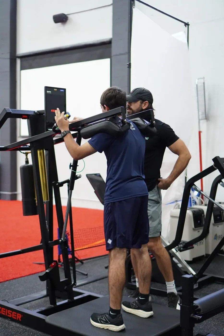 A coach watching an athlete’s squat on the Keiser machine