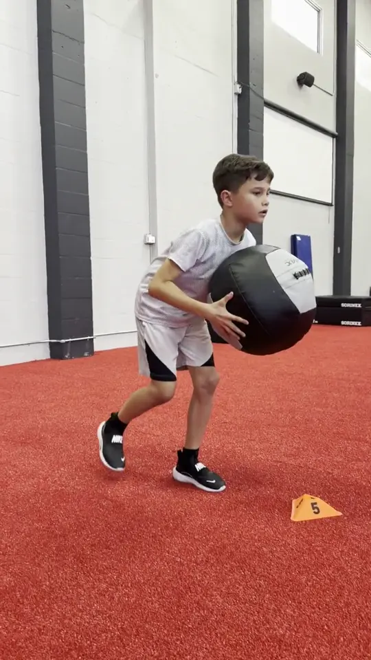 A young athlete carrying a medicine ball through a coordination drill on the turf